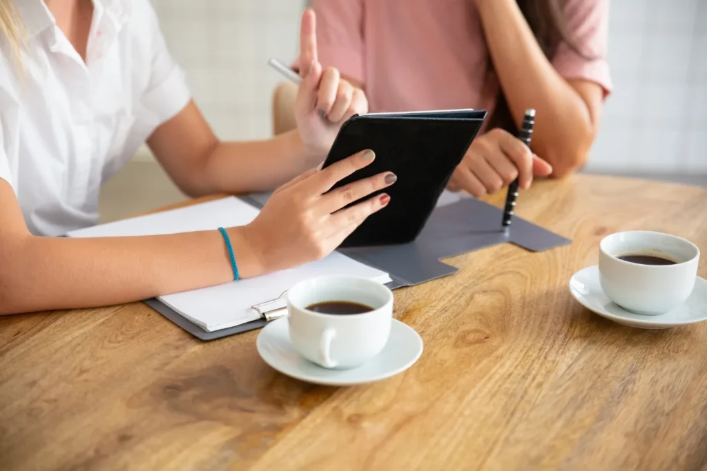 Due persone sedute a un tavolo di legno, inquadrate senza il volto: una cartellina con dei fogli, un tablet e due tazze di caffè.