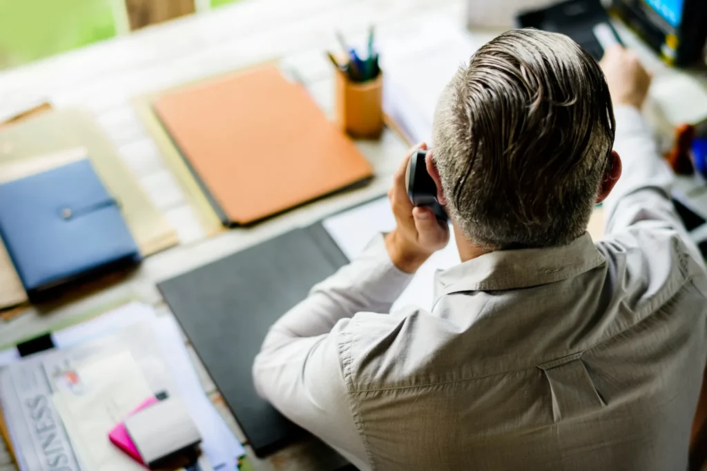 Un uomo ripreso di spalle che parla al telefono alla sua scrivania, fra cartelle e documenti.