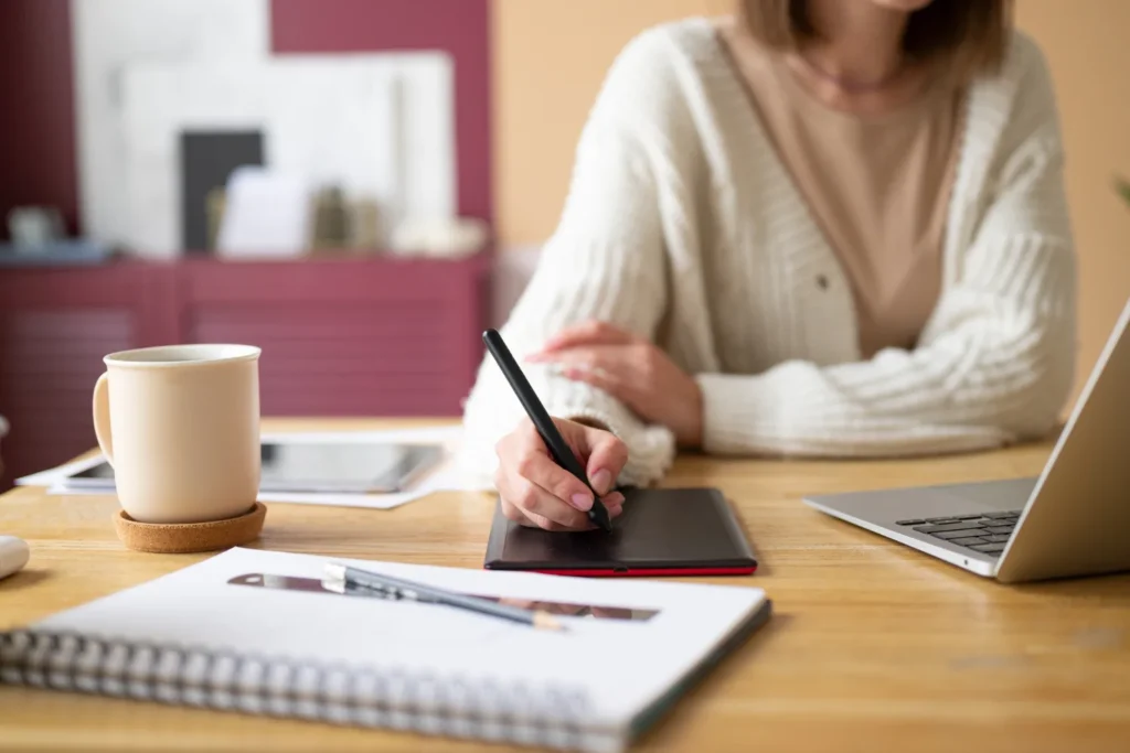 Una donna che scrive su un quaderno al tavolo di casa, accanto a una tazza e a un portatile.