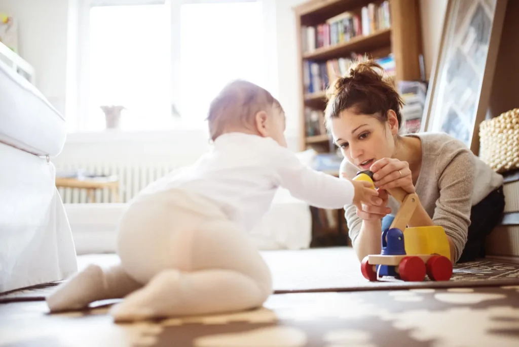 Una baby sitter che gioca sul tappeto con un bambino piccolo, in un soggiorno luminoso.